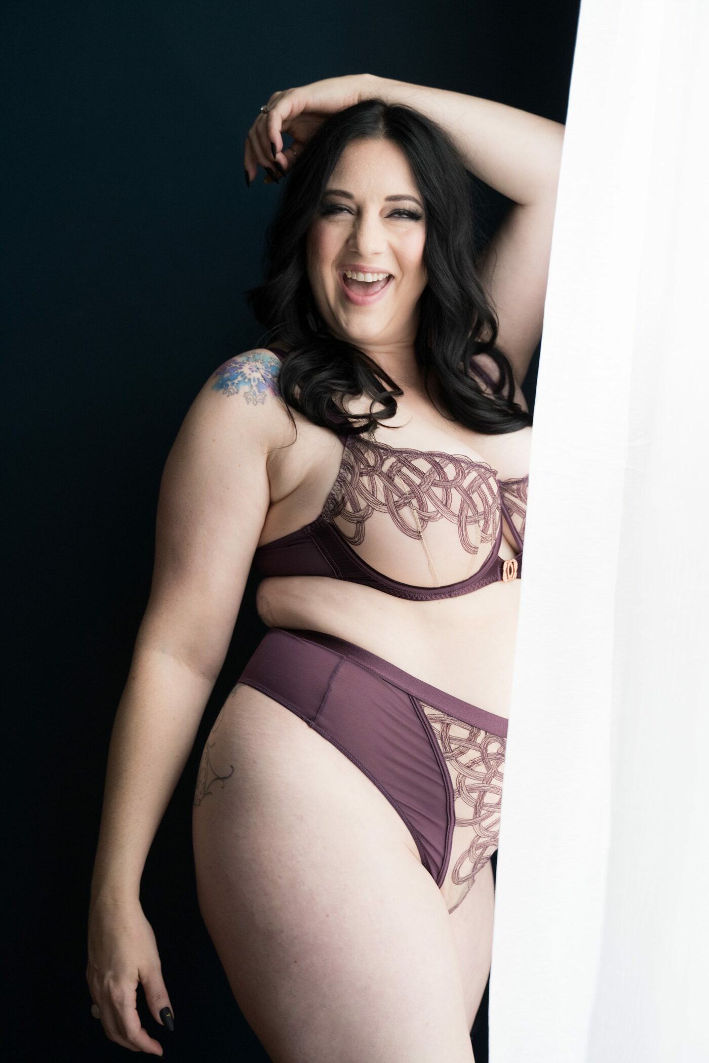 Playful portrait of woman in her 40s smiling, standing next to a window, wearing purple patterned bra and underwear.