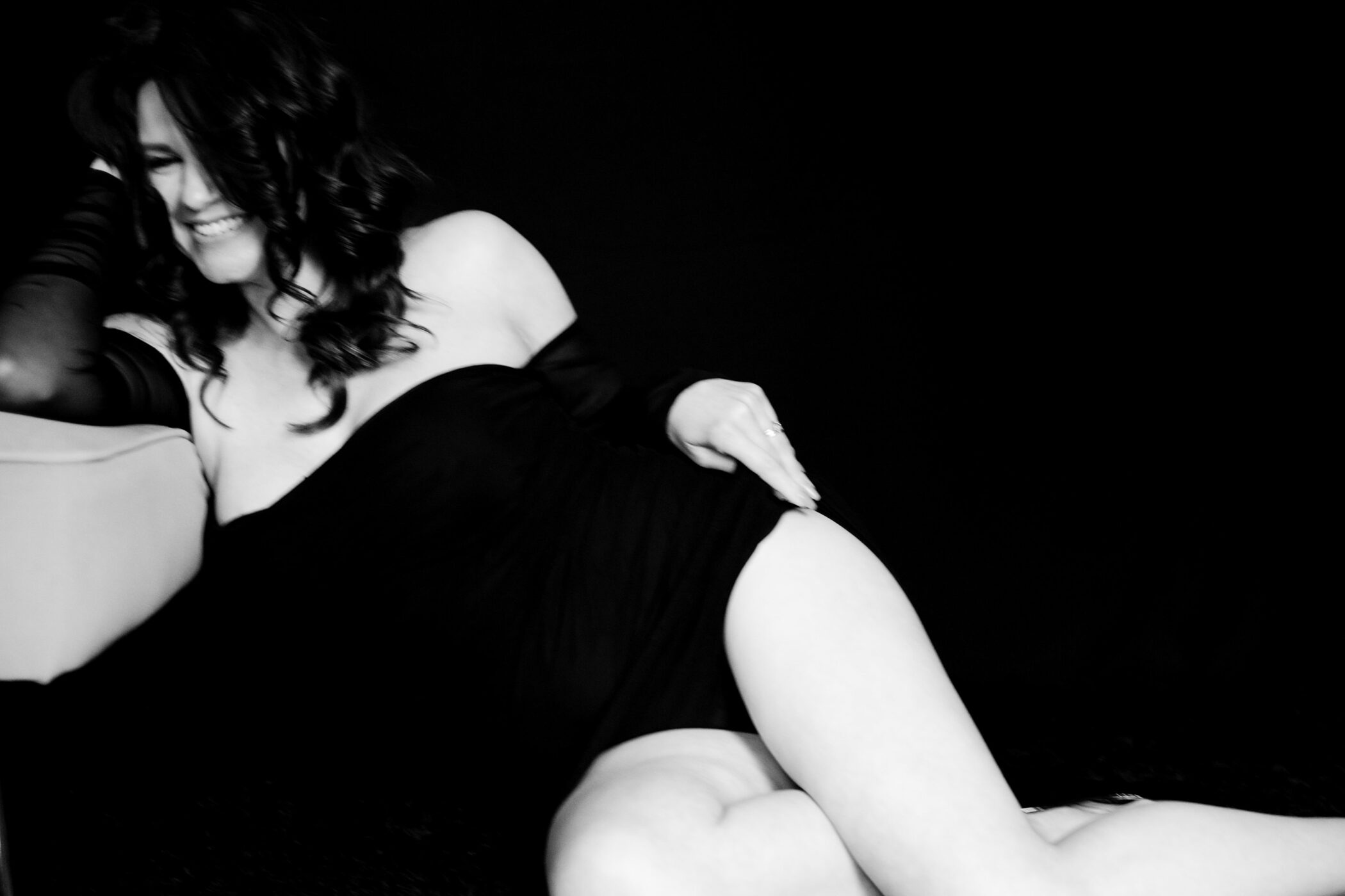 Moody black-and-white portrait of a woman in her 40s smiling touching her hip, wearing a black dress, seated against a black background with her elbow propped on a chair.