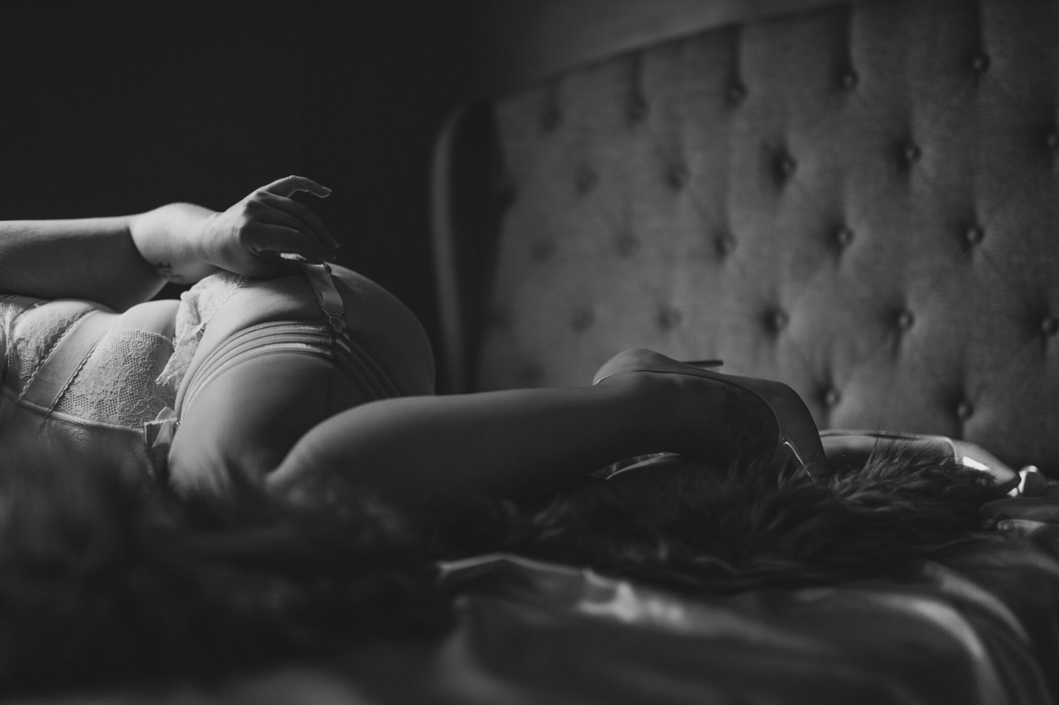 Close-up photo of woman touching garter belt, wearing high heels lying in-the-sheets, photographed in black-and-white.
