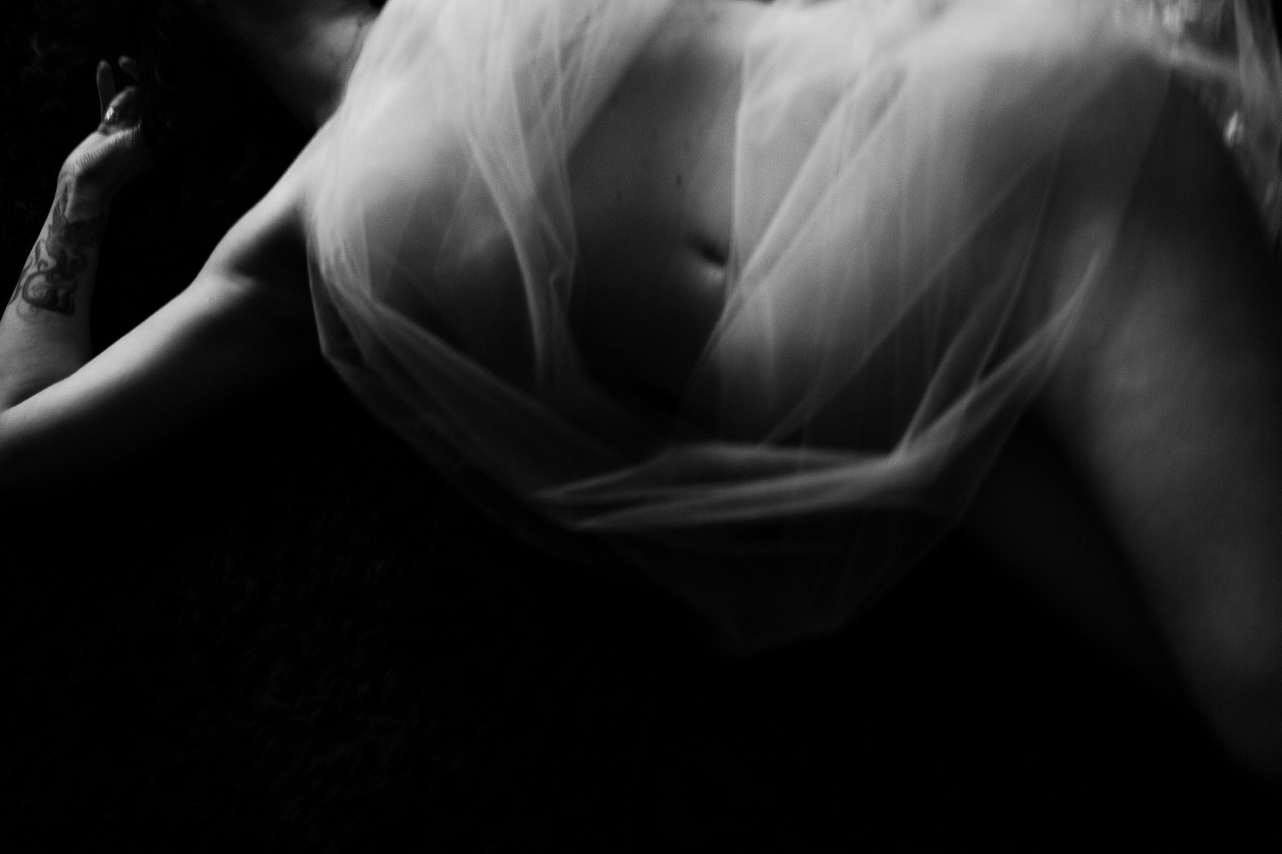 Moody black-and-white boudoir photo of woman lying against black background wrapped in tulle.