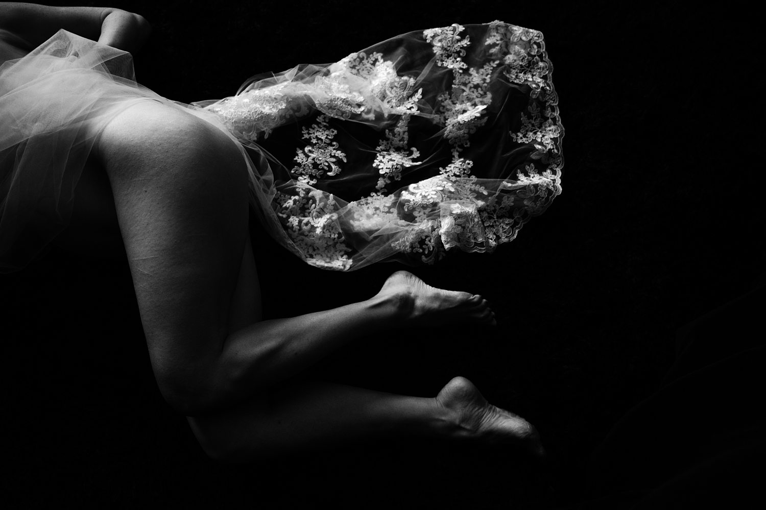 Artistic close-up of woman's legs draped in lace wedding veil, photographed in black-and-white.