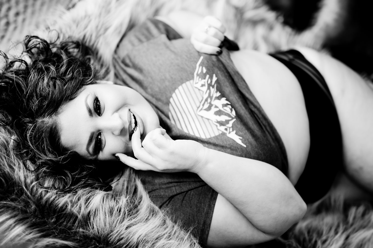 Cozy boudoir photo of woman wearing graphic t-shirt and black panties lying on a fur throw smiling at the camera.