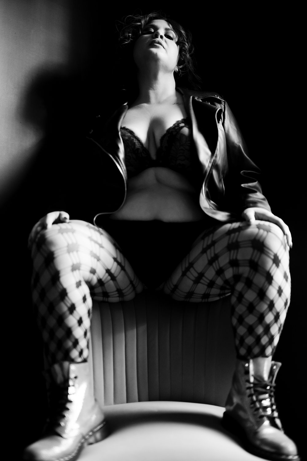 Edgy boudoir photo of woman wearing leather jacket and patterned stockings sitting atop a plush chair, photographed in black-and-white.