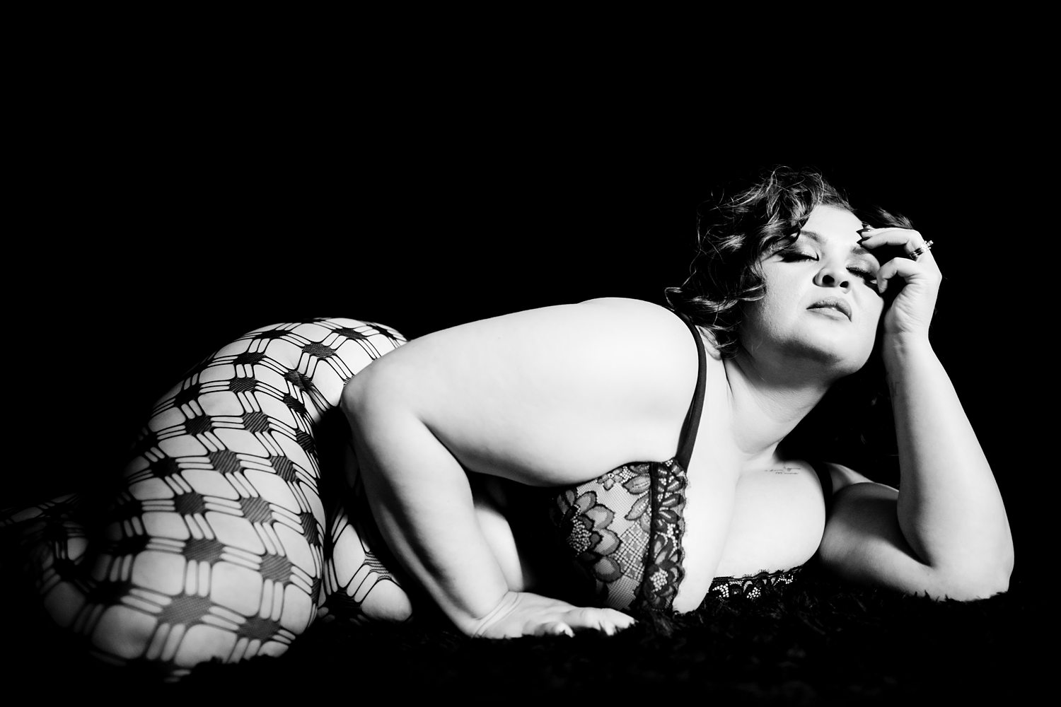Edgy photo of woman in patterned stockings and lace bra lying against a black background, photographed in black-and-white.