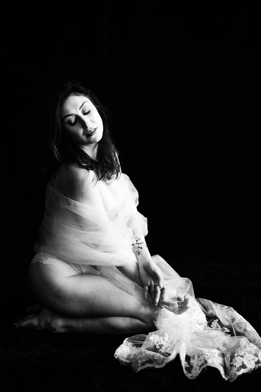 Editorial bridal portrait of woman wrapped in veil, kneeling against black background during artistic boudoir session.