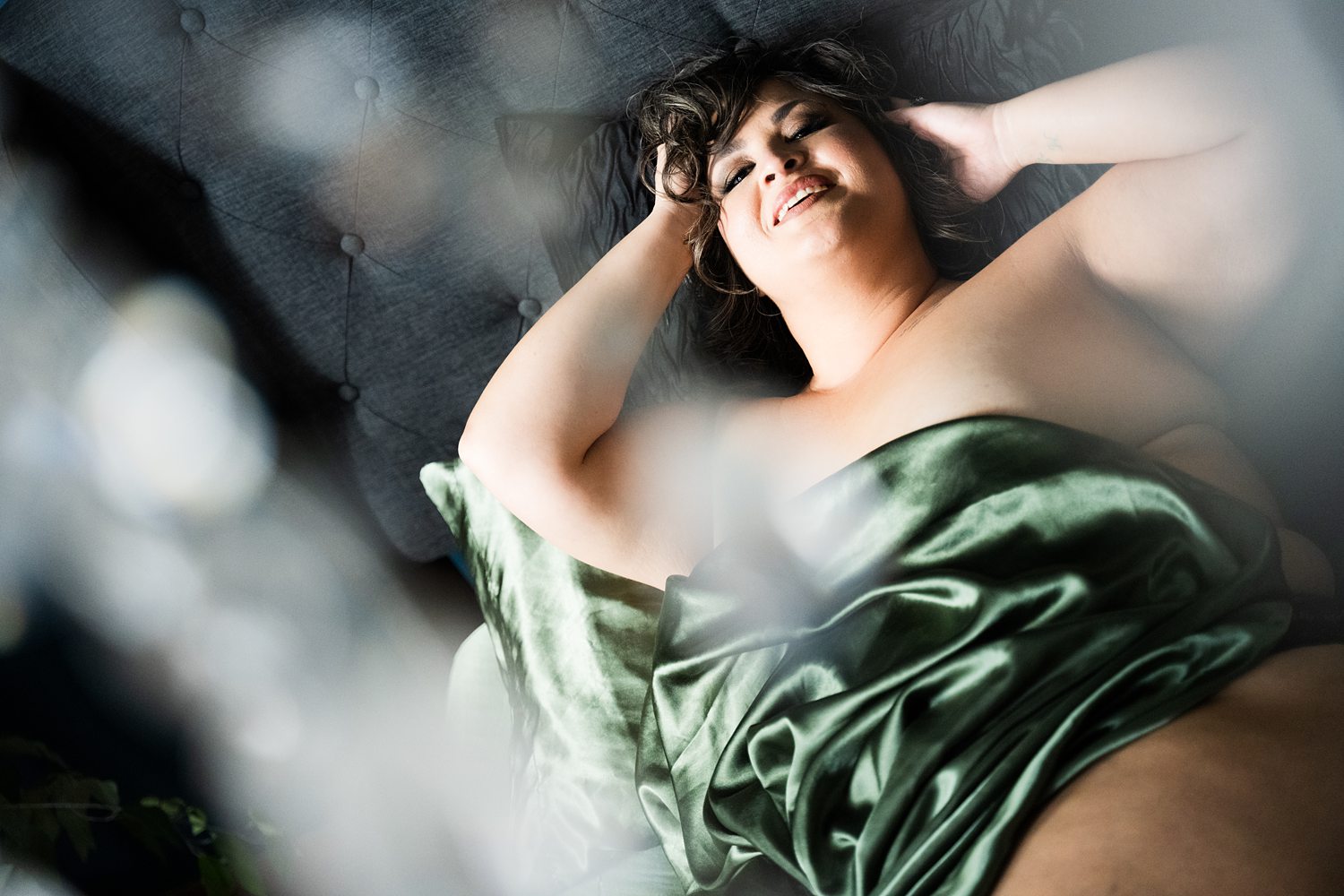 Flirtatious photo of woman lying on bed wrapped in green sheets smiling at the camera during soulful boudoir session.