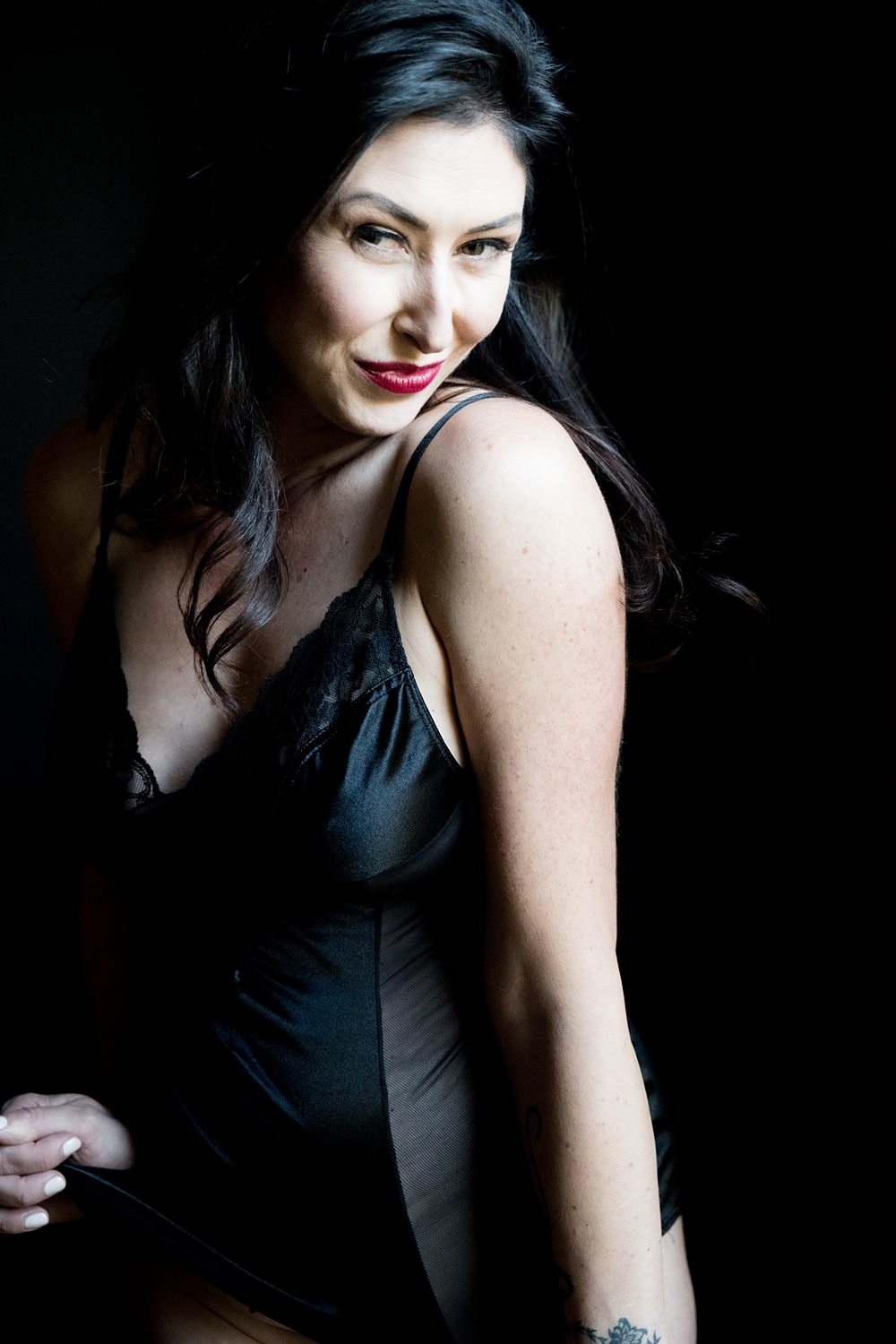 Flirtatious photo of woman wearing black lingerie slip confidently smiling at the camera.