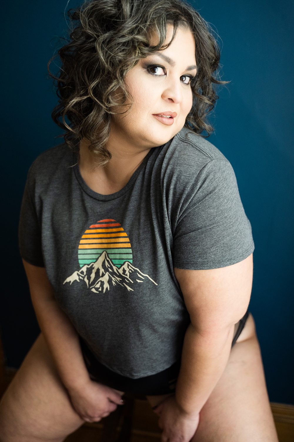 Portrait of woman wearing graphic t-shirt and black underwear grinning at the camera against a blue background.