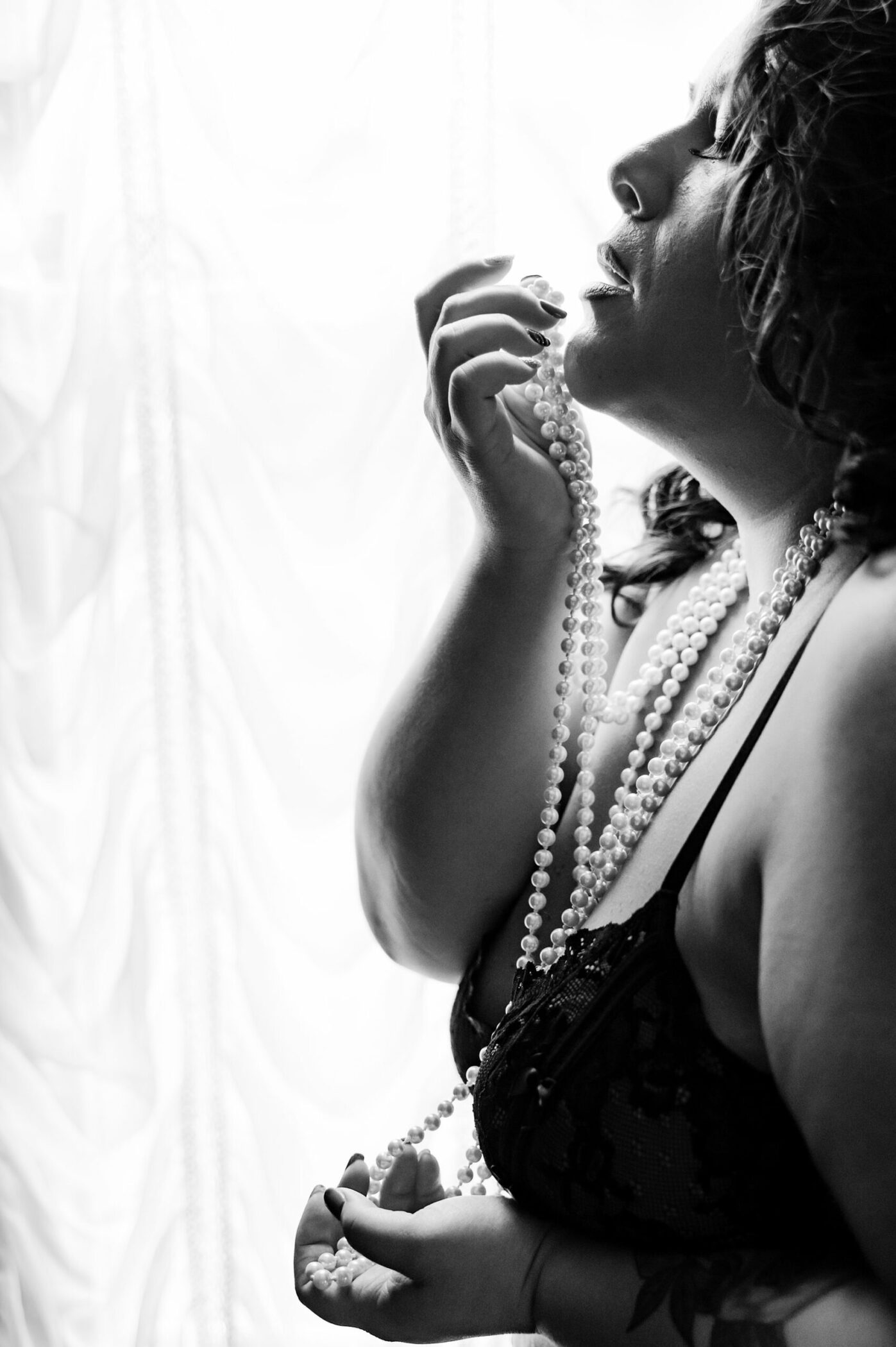 Sensual photo of woman wearing black lingerie and multiple strands of pearls standing in front of window.