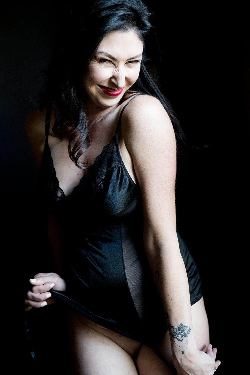 Woman gently pulling up her black lingerie slip, smiling at the camera.