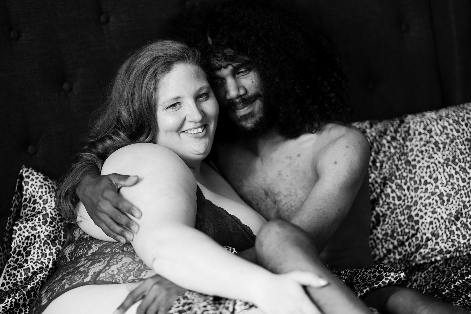 Smiling couple wrapped in each-others embrace lying on leopard print sheets during couples boudoir session.