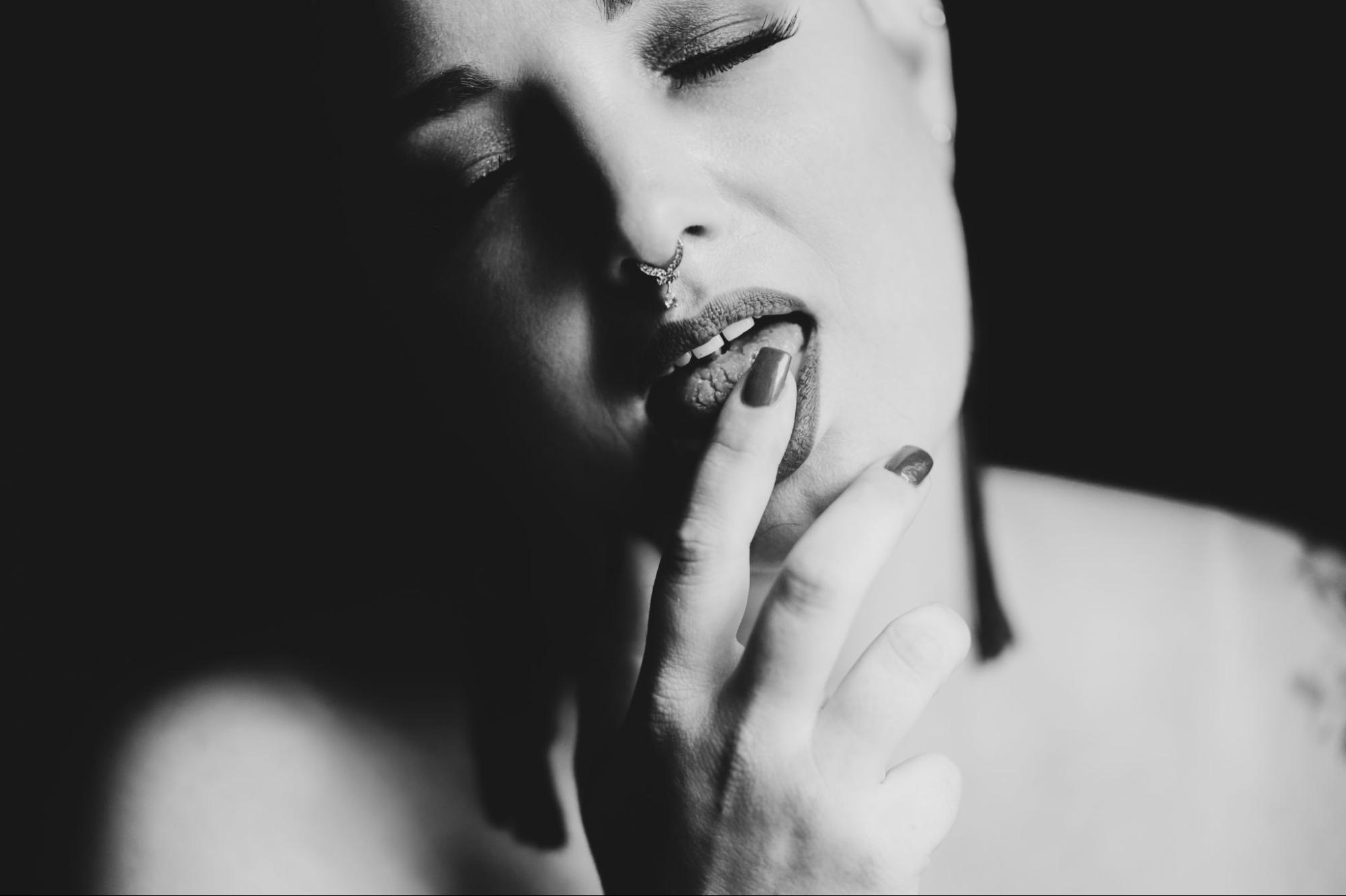 Erotic close-up of woman with septum piercing touching her finger to her tongue during artistic boudoir session.