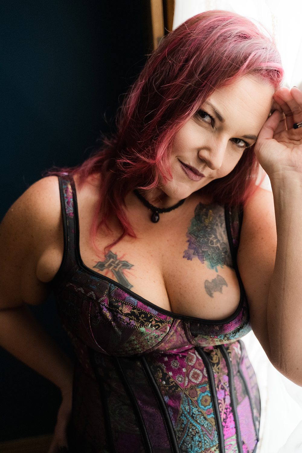 Boudoir photo of woman in purple patterned corset smiling at the camera leaning against a window.