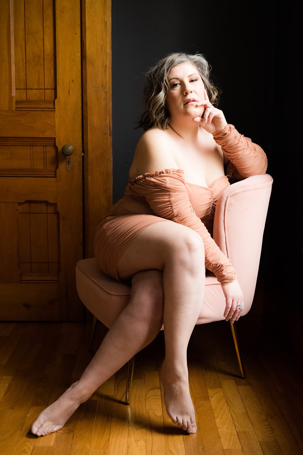 Boudoir photo of woman sitting on plush pink chair wearing blush colored dress looking confidently at the camera.