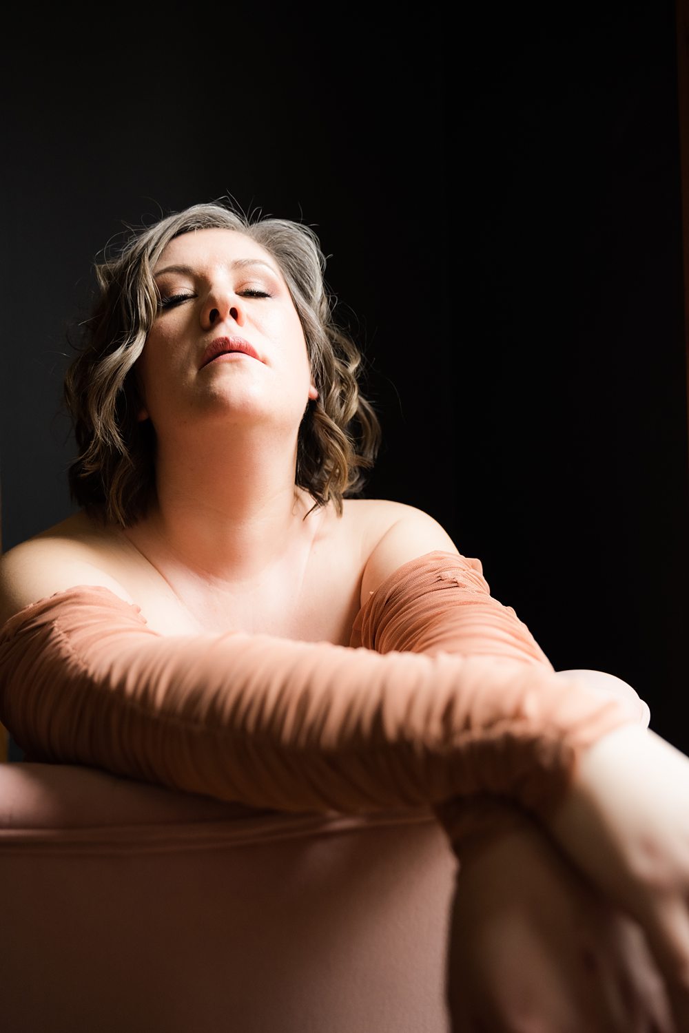 Emotive boudoir portrait of woman wearing a long sleeve dress sitting on a plush chair against a black backdrop.