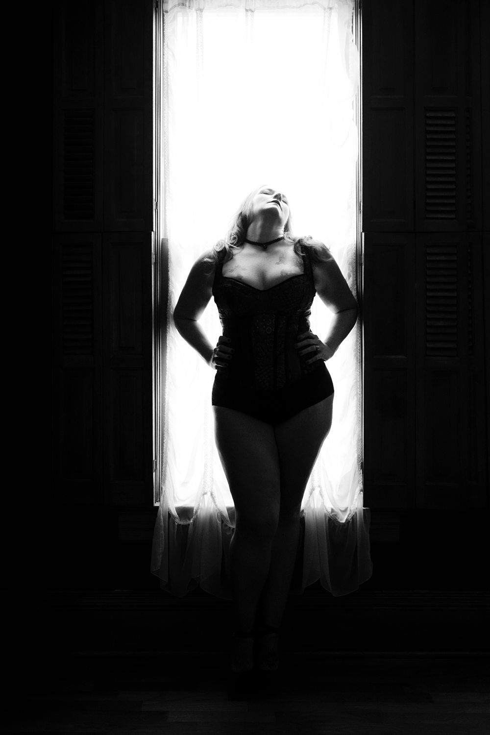 Soulful boudoir photo of a woman standing tall in front of a window, photographed in black-and-white.