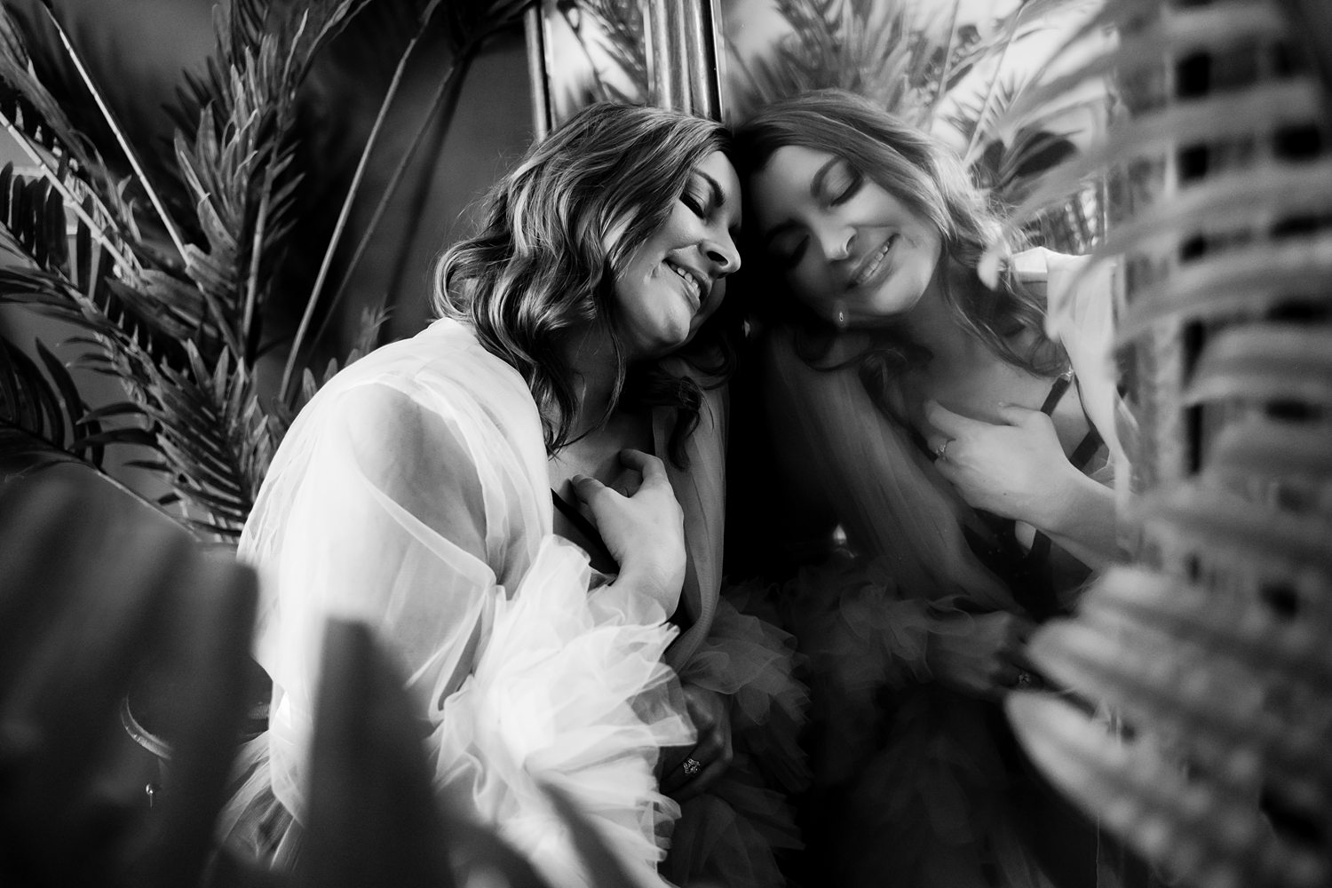 Black-and-white portrait of woman wearing tulle robe sitting at vintage vanity surrounded by plants during soulful boudoir session.