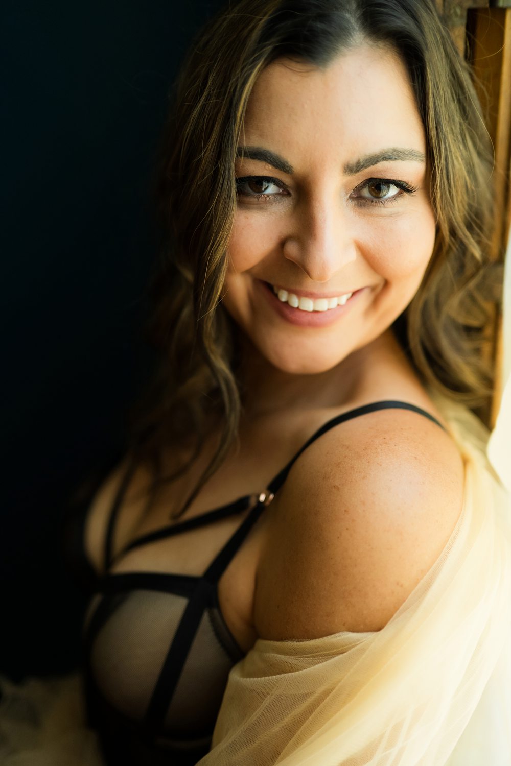 Close-up of woman smiling wearing black lingerie and tan tulle robe leaning against window frame.