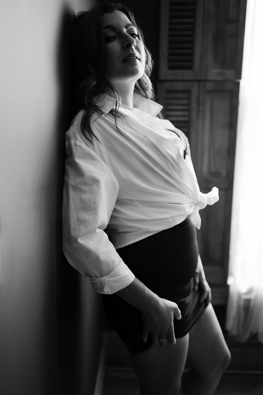 Woman wearing dress shirt confidently looking at camera leaning against wall, photographed in black and white.