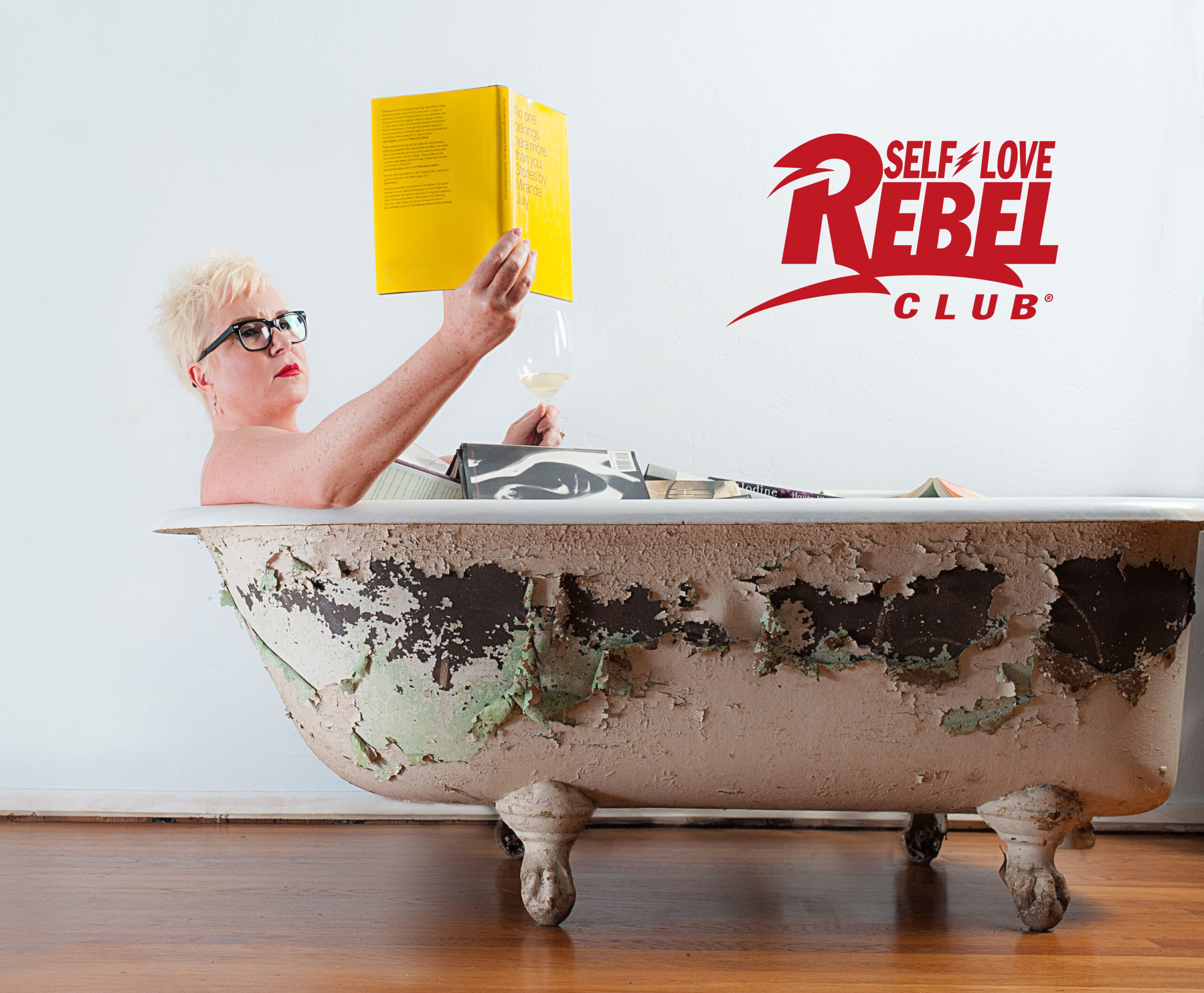 Woman sitting in a claw-foot tub confidently reading a book while holding a glass of wine.