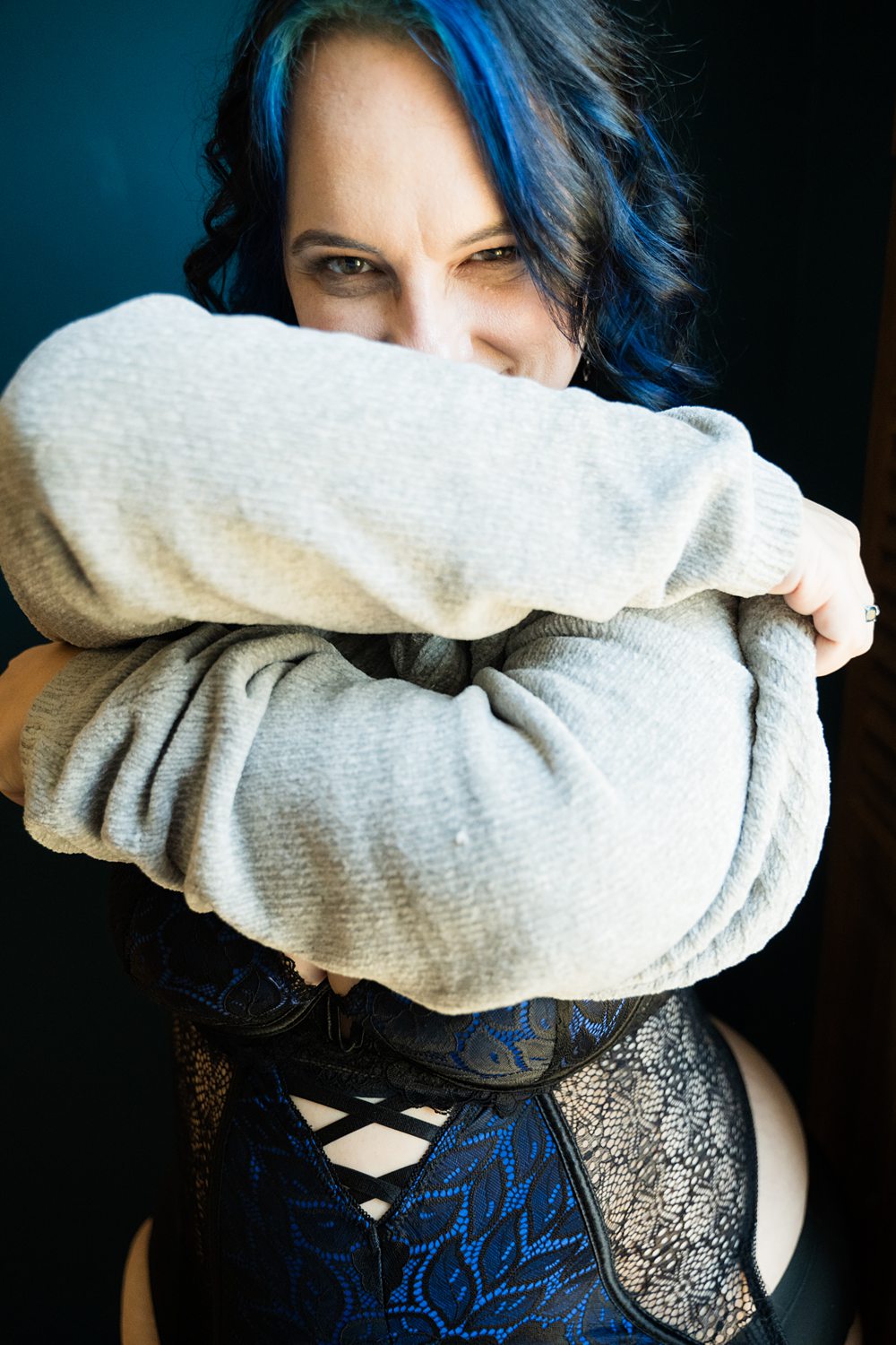 Playful boudoir photo of woman in black and blue lingerie taking off a grey sweater.
