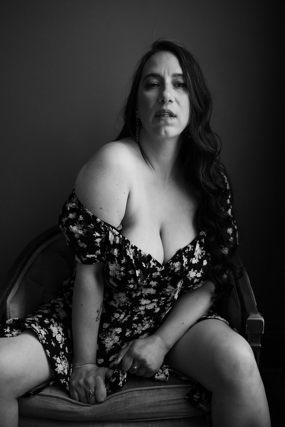 Alluring photo of a woman in her 40s wearing a low-cut dress, siting in a vintage chair during confidence building boudoir session.
