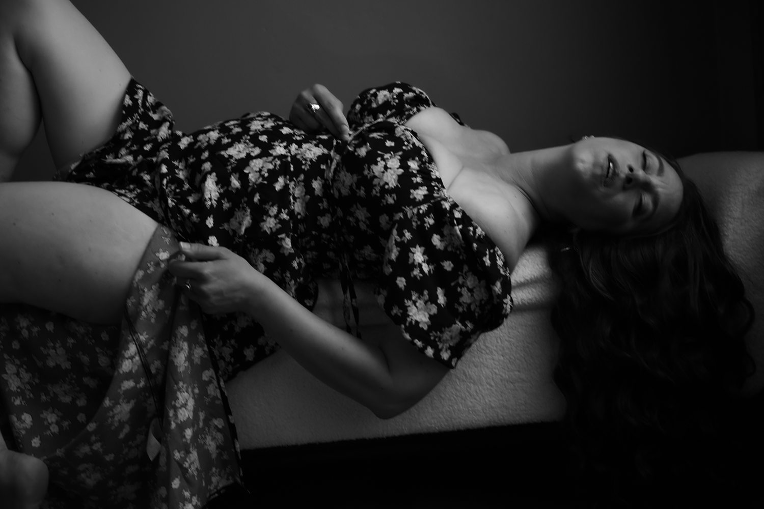 Black-and-white photo of woman lying on plush sofa, gently pulling up her dress during empowering boudoir session.