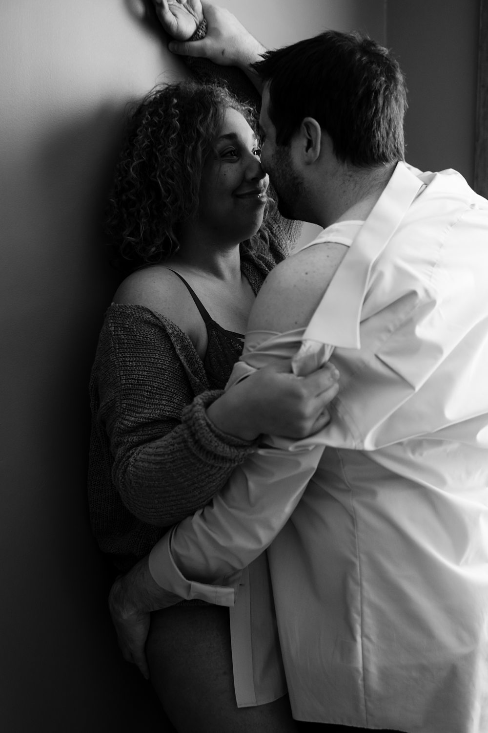 Black-and-white portrait of man wearing a dress shirt, leaning into a woman wearing lingerie against the wall.