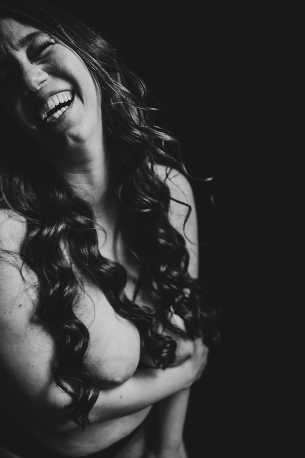 Playful black-and-white photo of a topless woman smiling joyfully at the camrea.