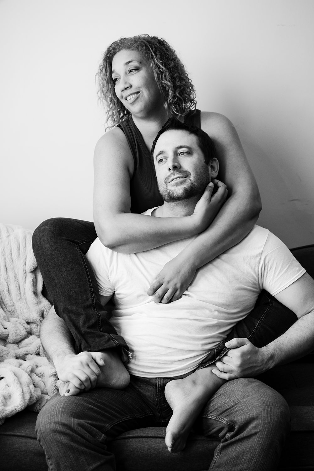 Playful black-and-white photo of a woman sitting behind her partner with her arms and legs wrapped around him.